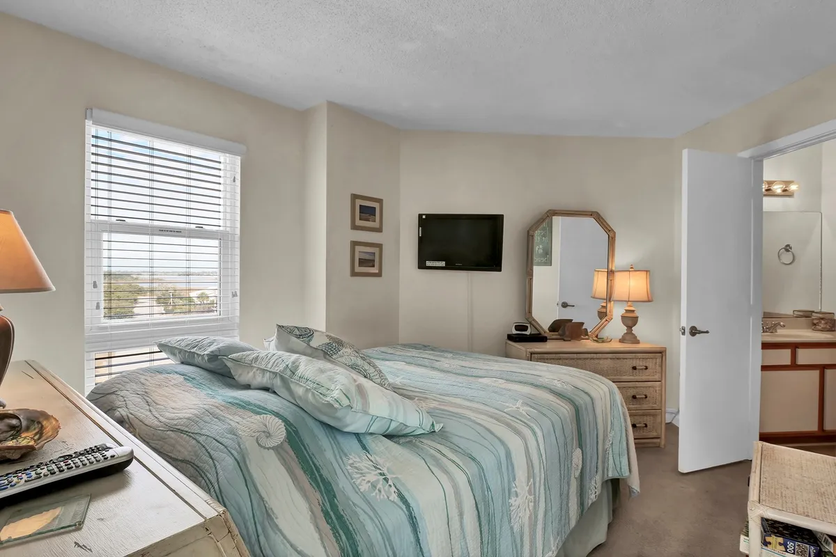 A tranquil master bedroom featuring a queen-sized bed adorned with a patterned quilt, a nightstand with a lamp, a wall-mounted TV, and a mirrored dresser. Sunlight filters softly into the room, creating a serene and restful atmosphere.