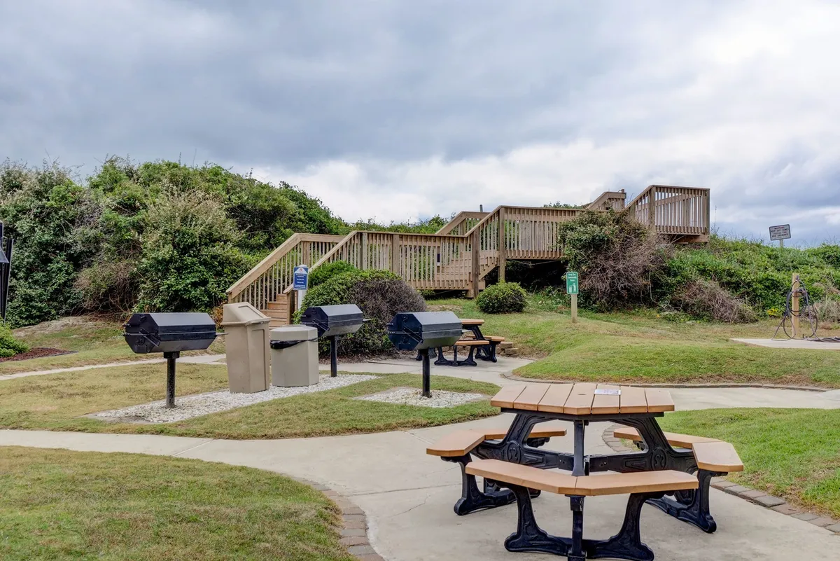 A relaxed communal area with picnic tables, grill stations, and a wooden staircase leading to nearby paths. The scene suggests gatherings, barbecues, and opportunities for socializing outdoors.