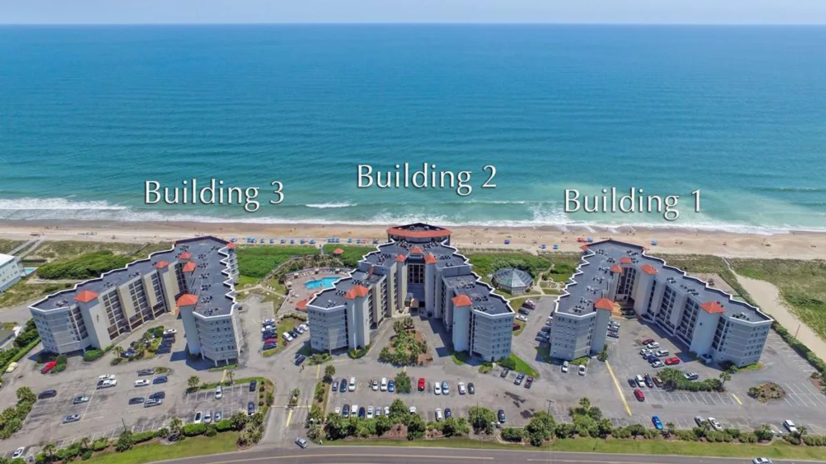 An exterior view of three resort buildings set around a sparkling swimming pool, all located beachfront. The ocean is visible beyond the pool, and the area exudes a relaxed, resort-style atmosphere ideal for sunbathing, swimming, or romantic evening walks by the shore.