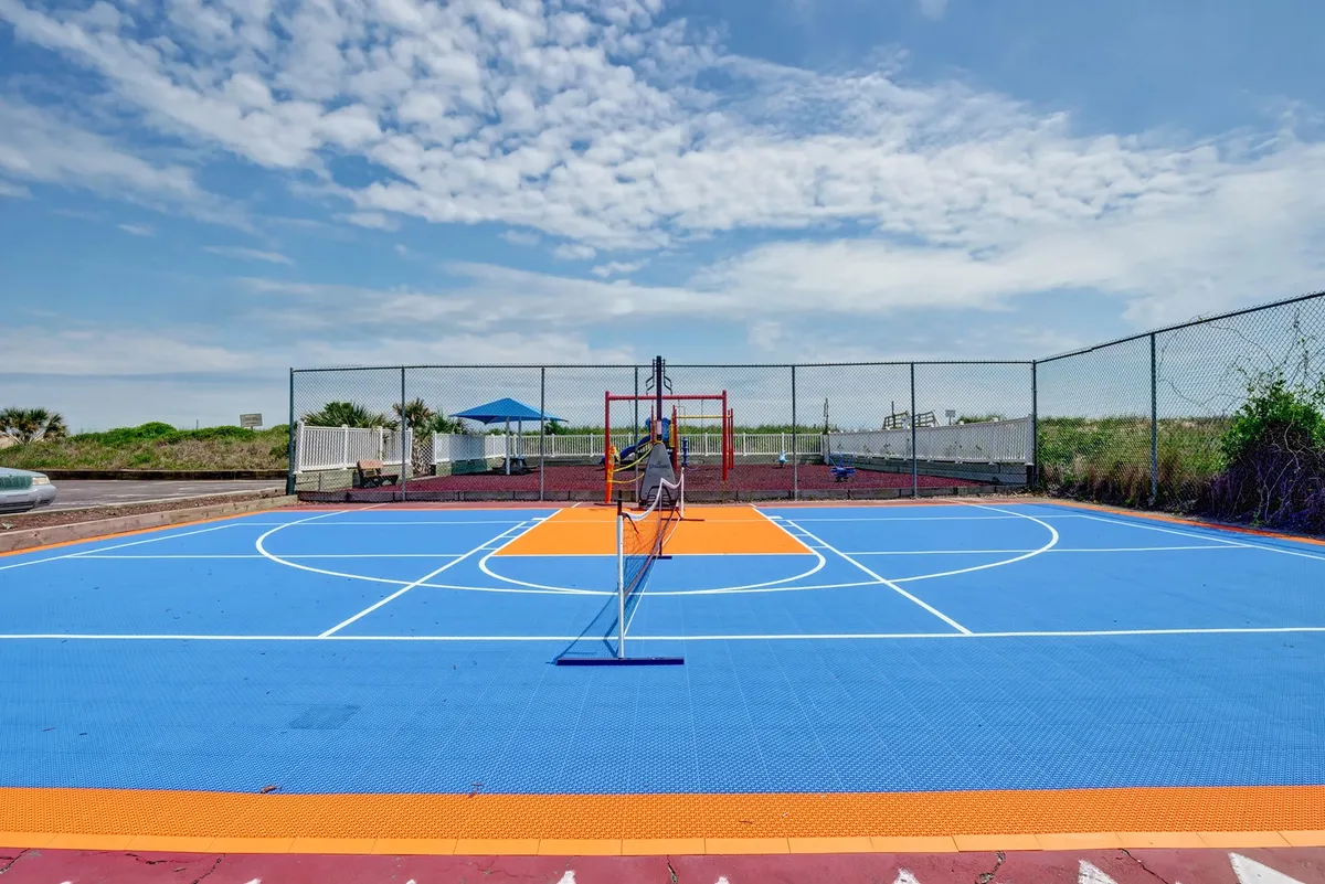 A vibrant exterior shot capturing the on-site playground, basketball court, and tennis net under a bright blue sky. Children are playing on the colorful playground equipment, while older kids and adults are engaged in basketball and tennis games. The surrounding greenery and open atmosphere evoke active, energetic days full of laughter and friendly competition.