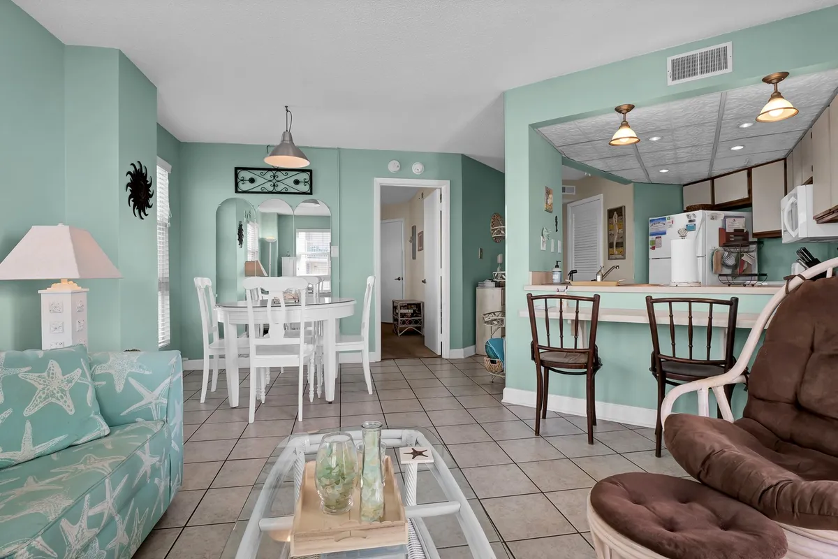 A bright interior featuring a starfish-patterned couch, a dining table, a kitchen bar, and a decorative mirror. The room is filled with natural light, and the open layout connects the living and dining areas with the kitchen.