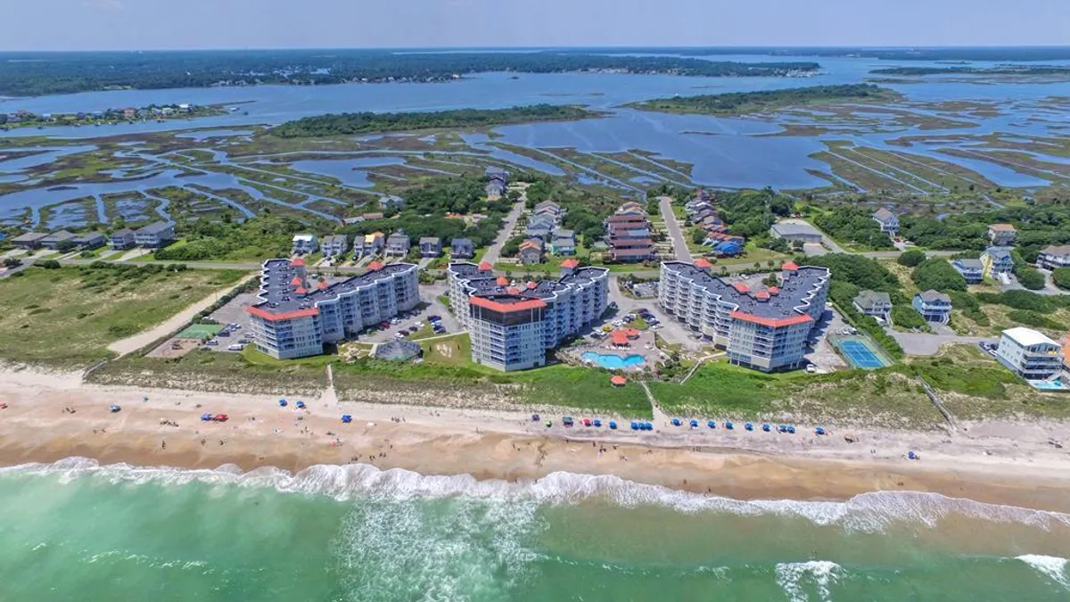 An aerial perspective of the beachfront property shows multiple resort buildings with a large swimming pool situated beside the sandy shoreline. Beach umbrellas dot the sand, with families relaxing and children playing nearby. The expansive pool area is clearly visible, symbolizing the newly updated and inviting saltwater pool mentioned in the text.