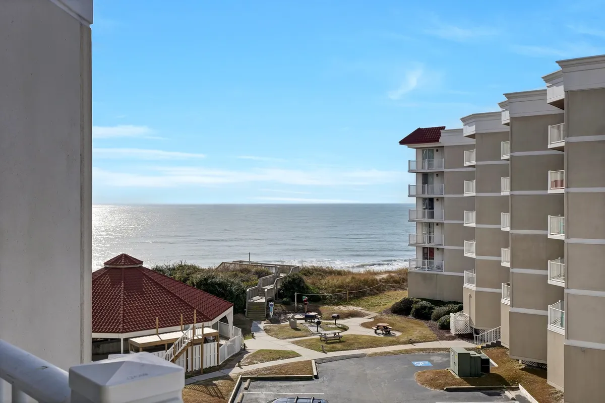The image showcases a stunning panoramic view of the North Topsail Beach shoreline, with sparkling ocean waters stretching to the horizon. In the foreground, a grassy area with a charming gazebo invites guests to relax or share a meal, while the condo building stands nearby, hinting at easy access to all the amenities. The tranquil scene evokes a sense of peace, with opportunities for picnicking, sunbathing, and beachside strolls clearly visible.