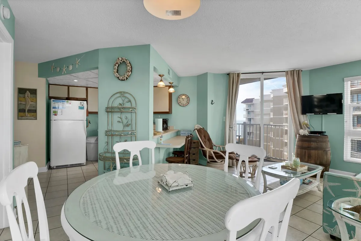 A bright interior space featuring a dining table and cozy couch, with a sliding door leading to a balcony. The view overlooks the ocean, emphasizing a tranquil setting for both work and relaxation. Natural light pours in, enhancing the inviting atmosphere.
