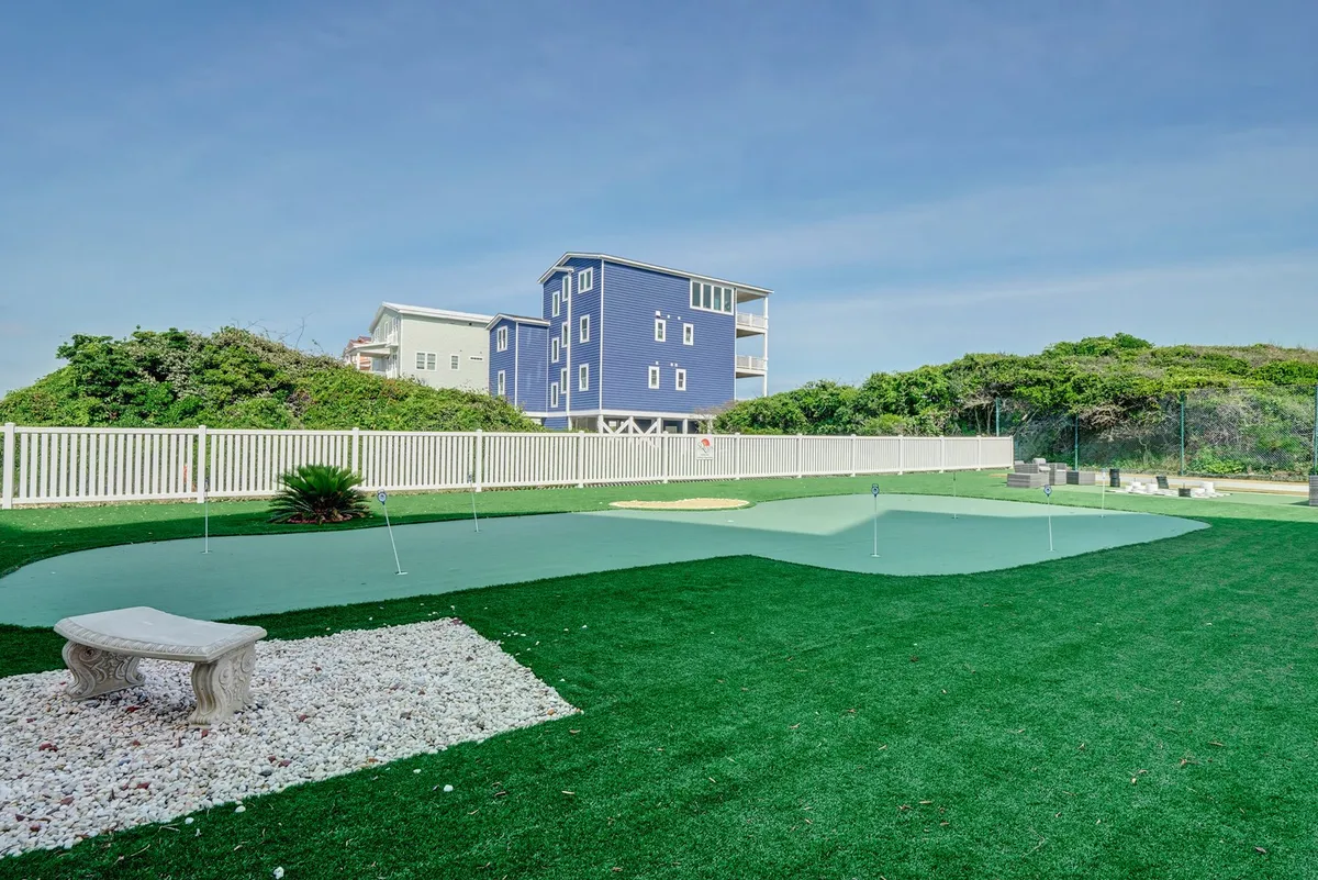 This image features a green putting green with a white stone pathway and a blue house in the background. A bench invites guests to relax, while the open space is ideal for group activities and casual golf practice.