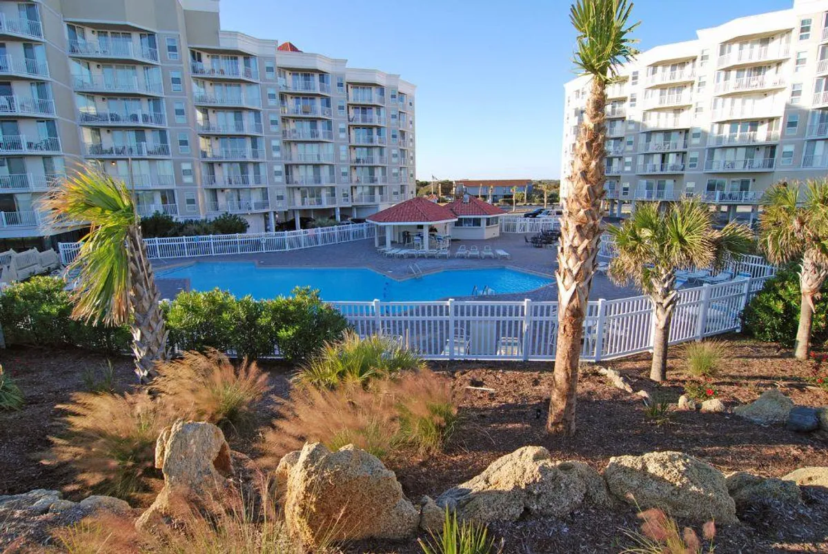 An inviting swimming pool surrounded by palm trees, landscaped gardens, and residential buildings. The pool is the focal point, with lounge areas suggesting spaces for relaxation and social gatherings. The lush, resort-style ambiance is enhanced by clear skies and vibrant greenery.