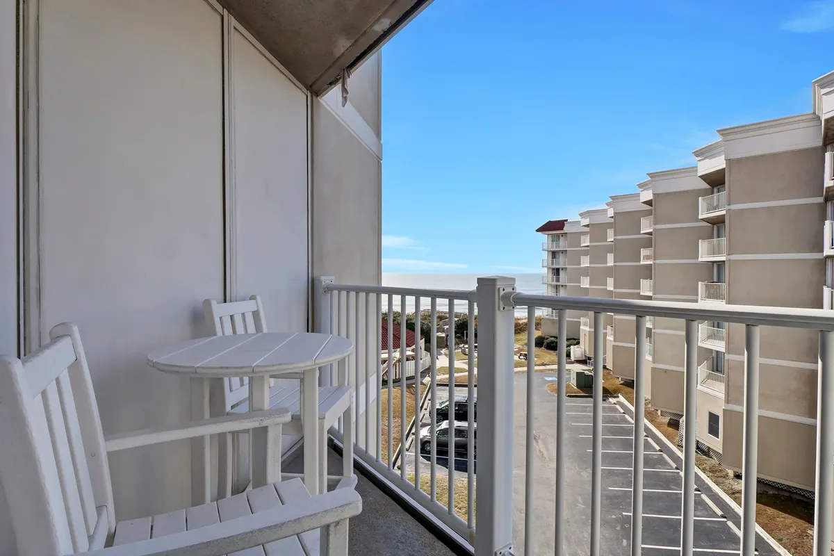 This image shows a private balcony with comfortable seating, offering an unobstructed view of the ocean. The scene captures a tranquil moment where the sun is either rising or setting, casting a warm glow over the water. The inclusion of neighboring buildings places the balcony within its beachfront context, enhancing the sense of a personal retreat connected to the larger coastal environment.